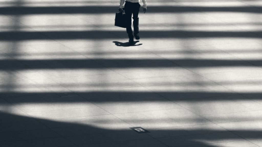 A lone figure crossing bands of light and shadow